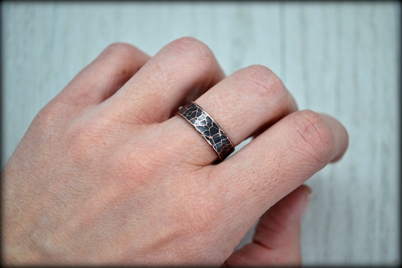 May include: A hammered copper ring with a dark, textured pattern. The ring is worn on a finger, showcasing its unique, handcrafted design. The band has a width of approximately 0.25 inches.