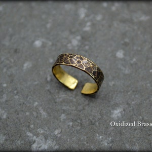 May include: An open-ended brass ring with an oxidized, textured band. The band features a dark brown, geometric pattern. The inner surface of the ring is a bright gold color. The text "Oxidized Brass" is visible.