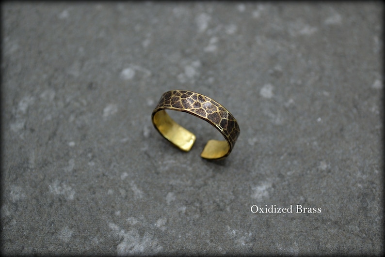 May include: An open-ended brass ring with an oxidized, textured band. The band features a dark brown, geometric pattern. The inner surface of the ring is a bright gold color. The text "Oxidized Brass" is visible.