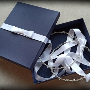 May include: A navy blue gift box with a white satin ribbon and bow. Inside, two delicate, beaded headbands with white ribbons are nestled. The headbands feature clear beads and small pearls, creating an elegant, bridal theme.