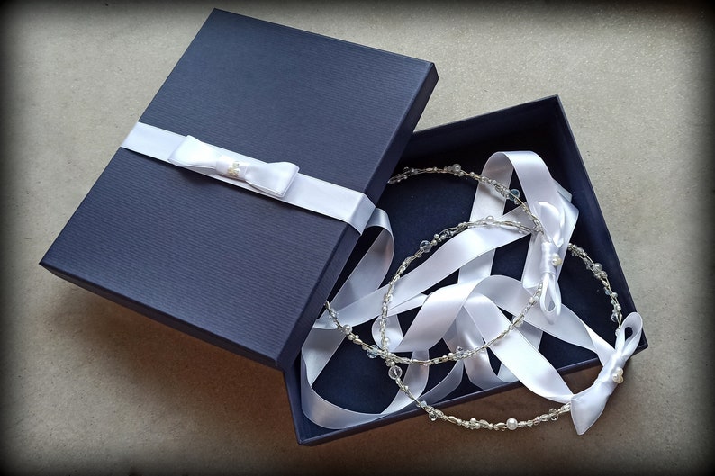 May include: A navy blue gift box with a white satin ribbon and bow. Inside, two delicate, beaded headbands with white ribbons are nestled. The headbands feature clear beads and small pearls, creating an elegant, bridal theme.