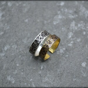 May include: Two adjustable rings with a textured, cracked pattern. One ring is silver with a black pattern, and the other is gold-toned with a brown pattern. The rings are displayed on a gray surface.
