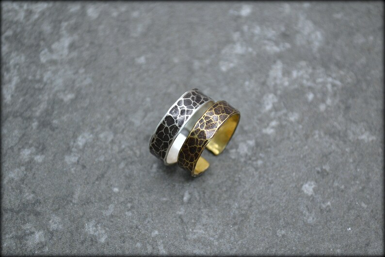 May include: Two adjustable rings with a textured, cracked pattern. One ring is silver with a black pattern, and the other is gold-toned with a brown pattern. The rings are displayed on a gray surface.