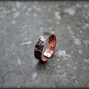 May include: A copper ring with a hammered texture and a dark patina. The ring has a band with a textured surface and a warm, reddish-brown color. The ring is open at the bottom.