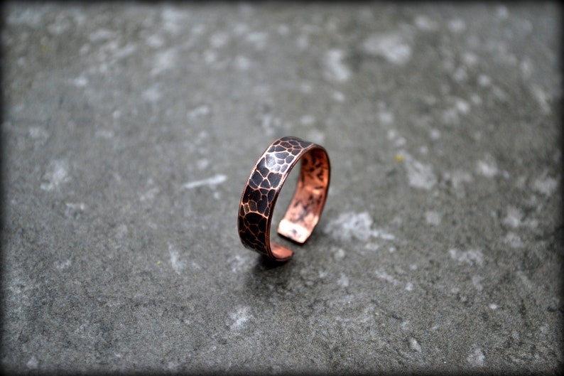 May include: A copper ring with a hammered texture and a dark patina. The ring has a band with a textured surface and a warm, reddish-brown color. The ring is open at the bottom.