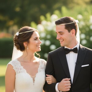 May include: A bride and groom are seen smiling at each other. The bride wears a white lace wedding dress and a gold headpiece. The groom is in a black tuxedo with a bow tie. They are outdoors in a garden setting.