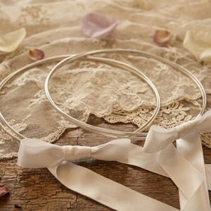 May include: Two silver wedding rings rest on a lace cloth, accompanied by a white ribbon and scattered rose petals. The rings are circular and simple, suggesting a focus on the ceremony.
