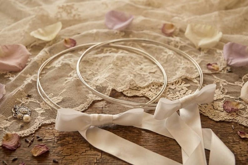 May include: Two silver wedding rings rest on a lace cloth, accompanied by a white ribbon and scattered rose petals. The rings are circular and simple, suggesting a focus on the ceremony.