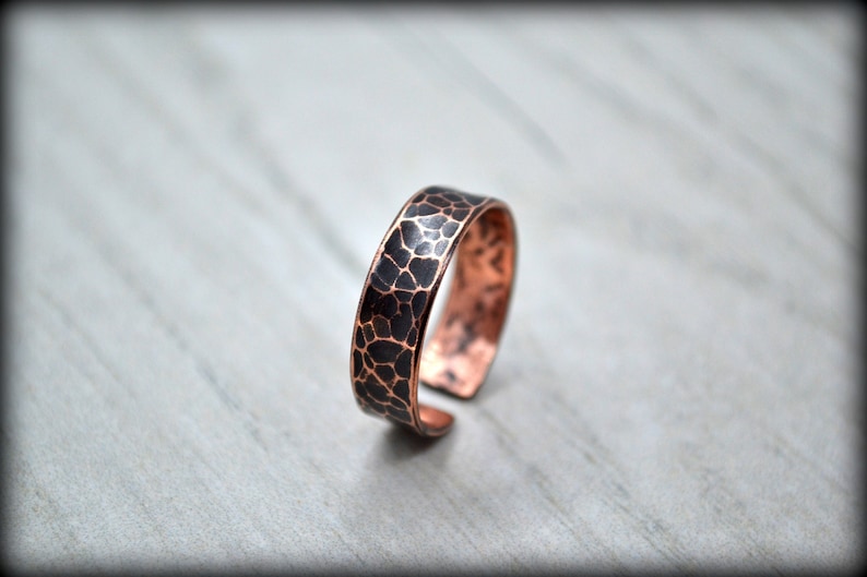 May include: A copper ring with a textured, dark pattern on the exterior. The ring has a hammered appearance and a warm, reddish-brown color. The ring is open at the bottom, allowing for size adjustment.