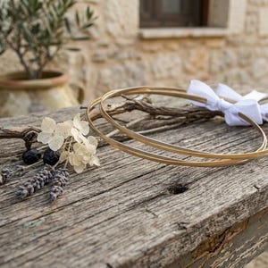 May include: Two gold wedding rings with a white ribbon bow rest on a weathered wooden surface, alongside dried flowers and sprigs. The rings are the focal point, suggesting a theme of marriage or commitment.