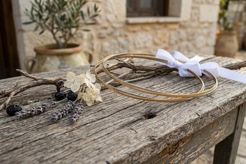May include: Two gold wedding rings with a white ribbon bow rest on a weathered wooden surface, alongside dried flowers and sprigs. The rings are the focal point, suggesting a theme of marriage or commitment.