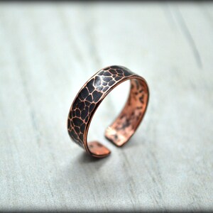 May include: A copper-colored ring with a textured, dark pattern on the top surface. The open-ended ring is displayed on a light gray surface. The ring's design features a unique, organic texture.