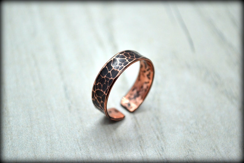 May include: A copper-colored ring with a textured, dark pattern on the top surface. The open-ended ring is displayed on a light gray surface. The ring's design features a unique, organic texture.