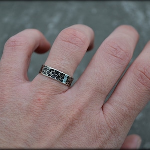 May include: A silver ring with a textured, black and silver pattern. The ring is worn on a finger, showcasing its unique design. The ring's surface has a mosaic-like appearance, adding to its visual appeal. The jewelry piece is a band style ring.
