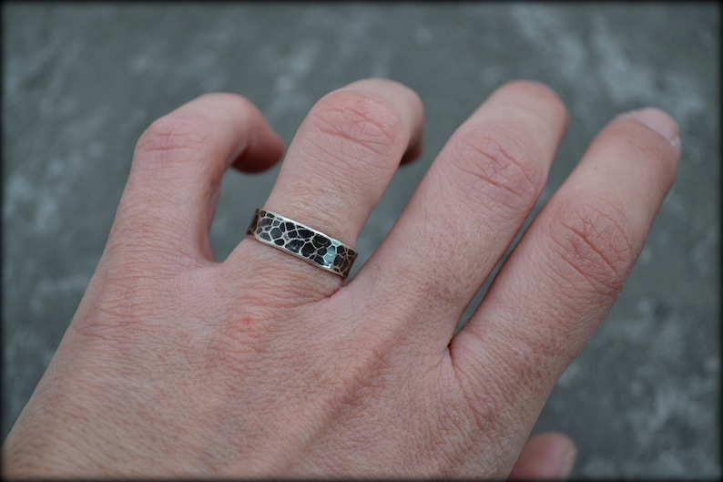 May include: A silver ring with a textured, black and silver pattern. The ring is worn on a finger, showcasing its unique design. The ring's surface has a mosaic-like appearance, adding to its visual appeal. The jewelry piece is a band style ring.