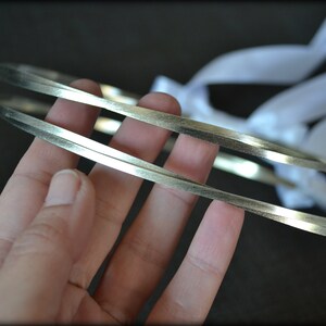 May include: Two silver-colored wedding crowns with a flat, rectangular design. White ribbons are attached to the crowns. The crowns are held in a hand against a dark background. The crowns are likely for a wedding ceremony.