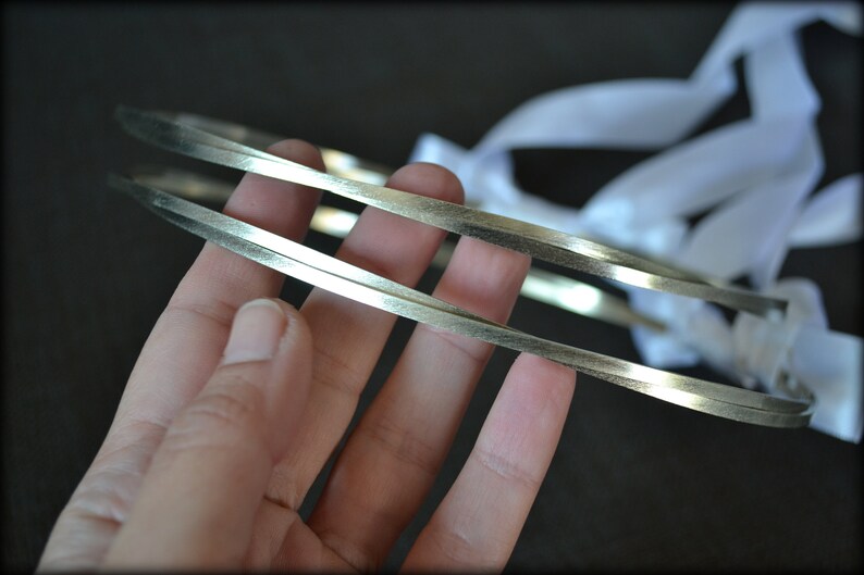 May include: Two silver-colored wedding crowns with a flat, rectangular design. White ribbons are attached to the crowns. The crowns are held in a hand against a dark background. The crowns are likely for a wedding ceremony.