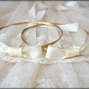 May include: Two gold-colored wedding crowns with ivory satin bows. The crowns are resting on a white lace and tulle surface. The bows are tied in a classic bow shape, adding a touch of elegance.