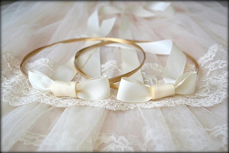 May include: Two gold-colored wedding crowns with ivory satin bows. The crowns are resting on a white lace and tulle surface. The bows are tied in a classic bow shape, adding a touch of elegance.
