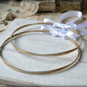May include: Two gold-toned wedding hoops with white satin bows, resting on a distressed white wooden surface. The hoops are thin and elegant, with a subtle shine. Vintage paper and lace add to the romantic aesthetic.