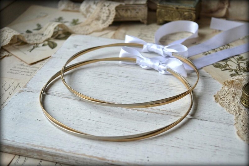 May include: Two gold-toned wedding hoops with white satin bows, resting on a distressed white wooden surface. The hoops are thin and elegant, with a subtle shine. Vintage paper and lace add to the romantic aesthetic.