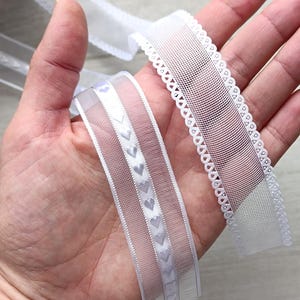 May include: Two white ribbons are held in a hand. One ribbon has a scalloped edge, the other has a series of small heart shapes. The ribbons are made of a sheer material and are likely used for crafts or embellishments.