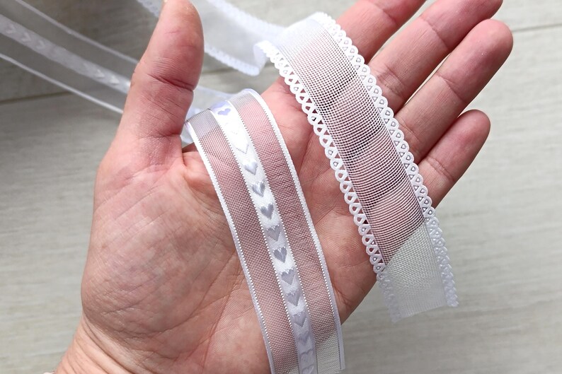 May include: Two white ribbons are held in a hand. One ribbon has a scalloped edge, the other has a series of small heart shapes. The ribbons are made of a sheer material and are likely used for crafts or embellishments.