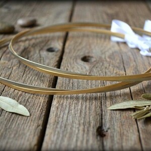 May include: Two gold wedding hoops with a white ribbon, resting on a rustic wooden surface. The hoops are thin and elegant, with a subtle shine. Decorative elements include small stones and leaves.