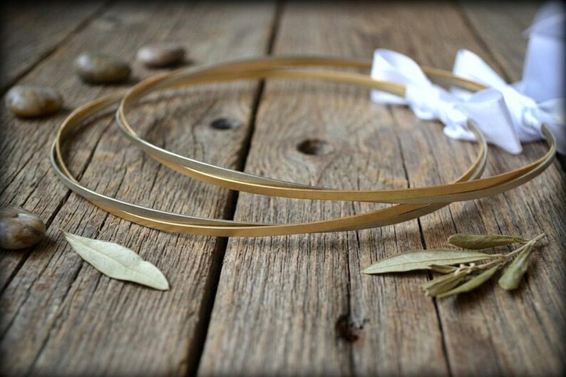 May include: Two gold wedding hoops with a white ribbon, resting on a rustic wooden surface. The hoops are thin and elegant, with a subtle shine. Decorative elements include small stones and leaves.