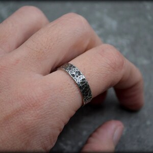 May include: A silver ring with a hammered texture, worn on a finger. The ring has a band with a dark, irregular pattern. The ring is a piece of jewelry, likely for everyday wear.