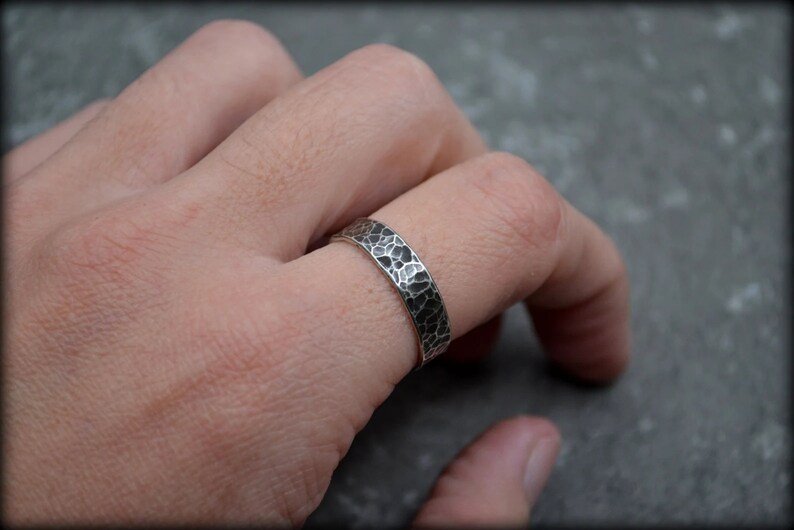 May include: A silver ring with a hammered texture, worn on a finger. The ring has a band with a dark, irregular pattern. The ring is a piece of jewelry, likely for everyday wear.