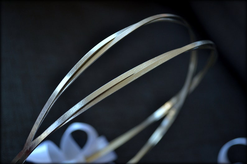 May include: Close-up of a silver-toned wedding crown with a delicate, intertwined design. The crown features a ribbon at the base and is set against a dark background, highlighting its elegant simplicity. Ideal for bridal wear.
