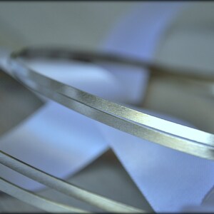 May include: Close-up of silver metal bands with a textured surface, layered over white satin ribbons. The bands are thin and have a slightly reflective quality, creating a sense of depth and elegance. The ribbons are soft and flowing.