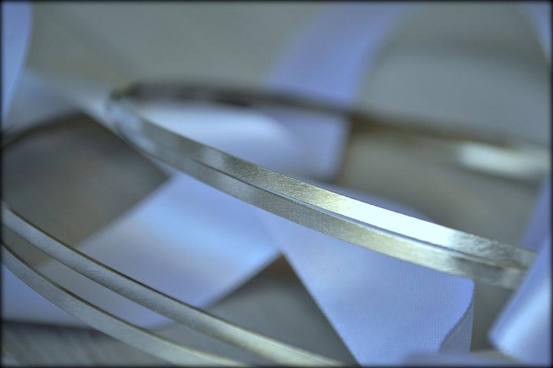 May include: Close-up of silver metal bands with a textured surface, layered over white satin ribbons. The bands are thin and have a slightly reflective quality, creating a sense of depth and elegance. The ribbons are soft and flowing.