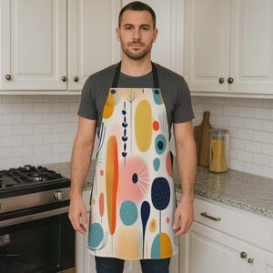 May include: An apron with a colorful abstract design featuring shapes in yellow, orange, pink, and blue on a cream background. The apron has black straps and is worn by a person in a kitchen setting.