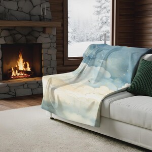 May include: A soft, watercolor-style throw blanket draped over a white sofa. The blanket features a light blue sky design with fluffy white clouds and gold star accents. A green pillow rests on the sofa. A fireplace with a roaring fire is in the background.