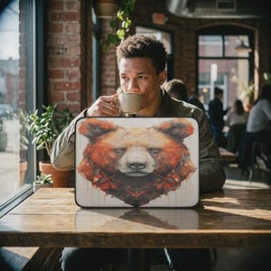 May include: A laptop sleeve featuring a watercolor illustration of a bear's face in shades of brown, red, and orange. The sleeve is rectangular and has a white background. The image is on a wooden table.