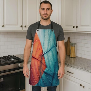 May include: An apron featuring a close-up of a leaf design in shades of orange and teal. The apron has a black neck strap and is worn by a person in a kitchen setting. The apron's design is a vibrant and artistic representation of nature.