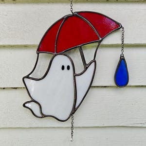 May include: A stained glass suncatcher featuring a white ghost holding a red umbrella. A single blue teardrop-shaped glass piece hangs from the edge of the umbrella by a small metal chain. The entire piece is outlined in dark metal solder.