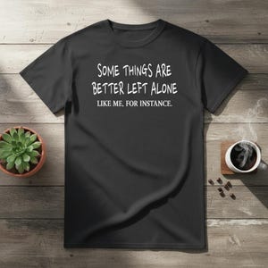 Puede incluir: Camiseta negra con el texto blanco "SOME THINGS ARE BETTER LEFT ALONE LIKE ME, FOR INSTANCE." Una pequeña maceta con suculentas y una taza de café con vapor son visibles sobre una superficie de madera.