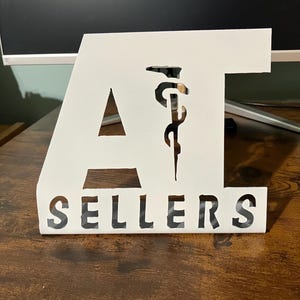 May include: White metal sign with the letters "AT" and the word "SELLERS" cut out. The "AT" includes a medical symbol. The sign is on a dark wooden surface.