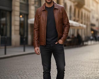 Puede incluir: Chaqueta de cuero marrón con cierre de cremallera, sobre una camiseta gris oscuro de cuello redondo. La persona lleva vaqueros negros y botas marrones. El fondo es una calle adoquinada.
