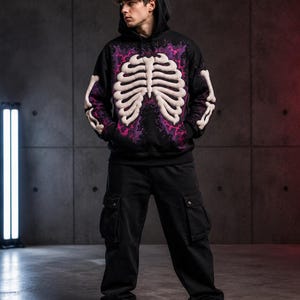 Puede incluir: Una sudadera negra con capucha que presenta un gráfico de caja torácica blanca en relieve en la parte delantera y las mangas, acentuado por un patrón de rayos morados y rosas. La capucha incluye dos pequeños cuernos morados, combinada con pantalones cargo negros y zapatillas blancas.