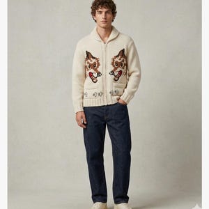 May include: Cream-colored zip-up cardigan sweater with two cartoon dog designs on the front. The sweater has a shawl collar, two front pockets, and a decorative pattern near the bottom. The person is wearing dark blue jeans and cream-colored sneakers.