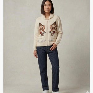 May include: Cream-colored zip-up cardigan sweater with a shawl collar and two front pockets. The sweater features a whimsical design of two brown and tan animal figures with red hearts. The person is wearing dark blue jeans and white sneakers.