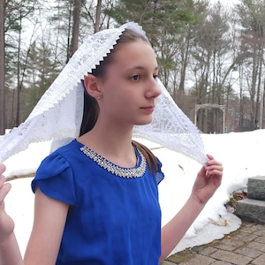 Puede incluir: Un velo de encaje blanco con borde festoneado está drapeado sobre la cabeza de una persona. La persona lleva una blusa azul real con un escote con cuentas. El fondo presenta árboles y nieve.