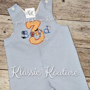 May include: A light blue and white gingham romper with the name "Boyd" and the number "3" embroidered in orange. Halloween-themed embroidery includes a spider web, bat, pumpkin, and ghost. The text "Klassic Kouture" is also visible.