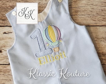 Könnte beinhalten: Ein blauer und weißer Gingham-Baby-Strampler mit einem Heißluftballon und der Zahl eins, die auf der Vorderseite gestickt sind. Der Name Elliott ist ebenfalls auf dem Strampler gestickt. Der Strampler wird von Klassic Kouture hergestellt.