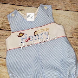 May include: A blue and white gingham baby romper with a white bib and embroidered cowboy theme. The bib has the name "Henry" and the text "Klassic Kouture".