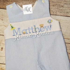 May include: A blue and white striped romper with a white embroidered bib. The bib has the text "Matthew Klassic Kouture" with a birthday hat, candle, and teddy bear illustrations.